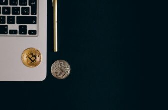 Close-up view of Bitcoin and Ethereum coins next to a laptop and pen on a black surface.
