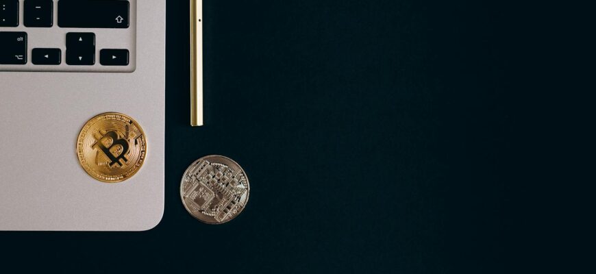 Close-up view of Bitcoin and Ethereum coins next to a laptop and pen on a black surface.